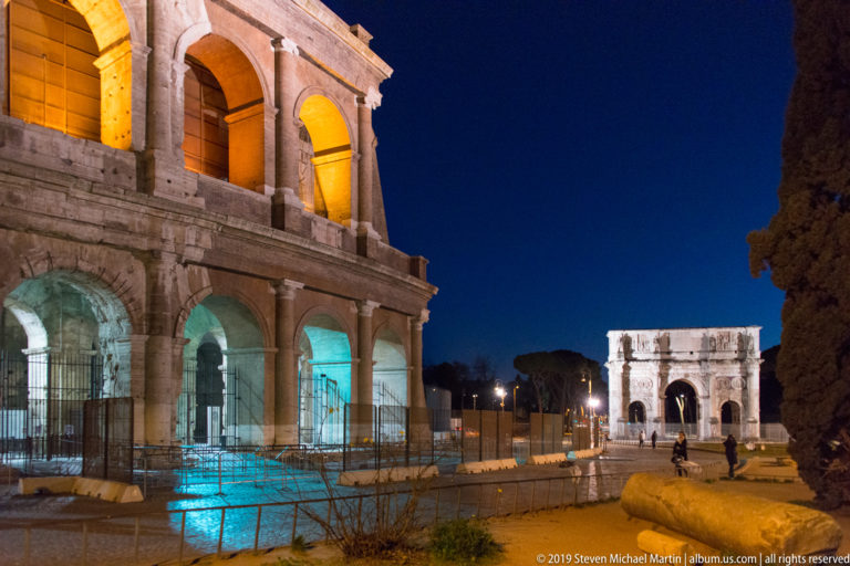 Night view with the Colosseum arcades at left and the illuminated Arch of Constantine farther down the walkway at right.