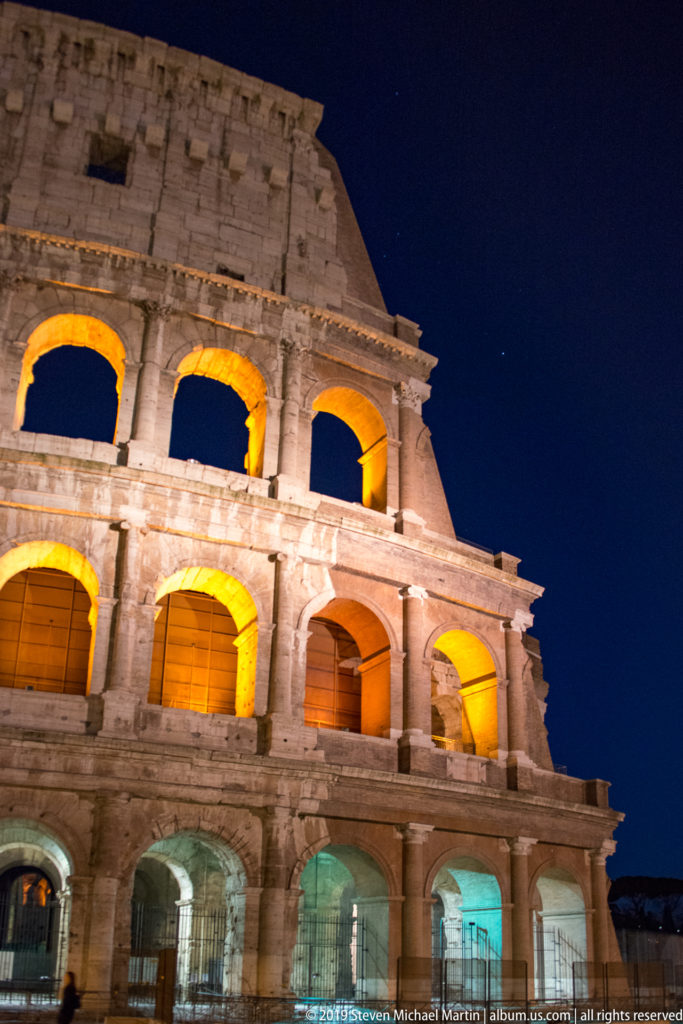 Upward night view of the Colosseum exterior with stacked illuminated arches rising diagonally against a dark blue sky.