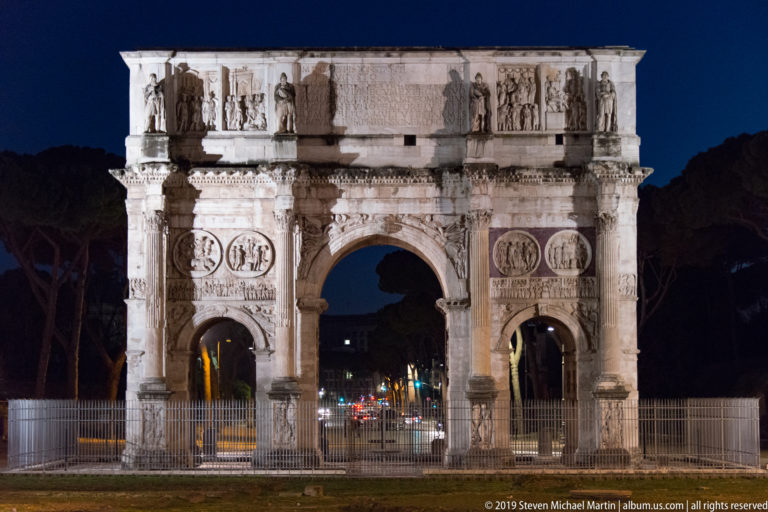 Floodlit Arch of Constantine at night, showing three archways, columns, sculpted roundels and relief panels, and the attic inscription.