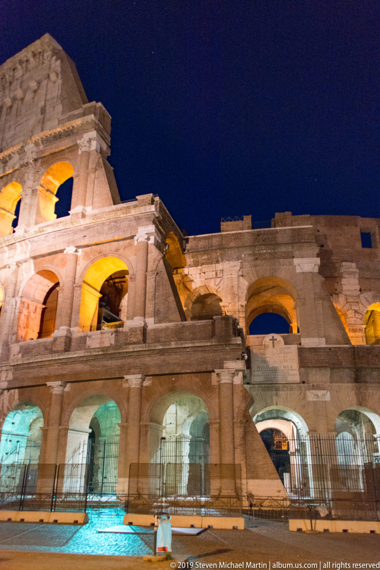 Close night view of the Colosseum’s lower and middle arcades, with illuminated openings and barriers along the pavement.
