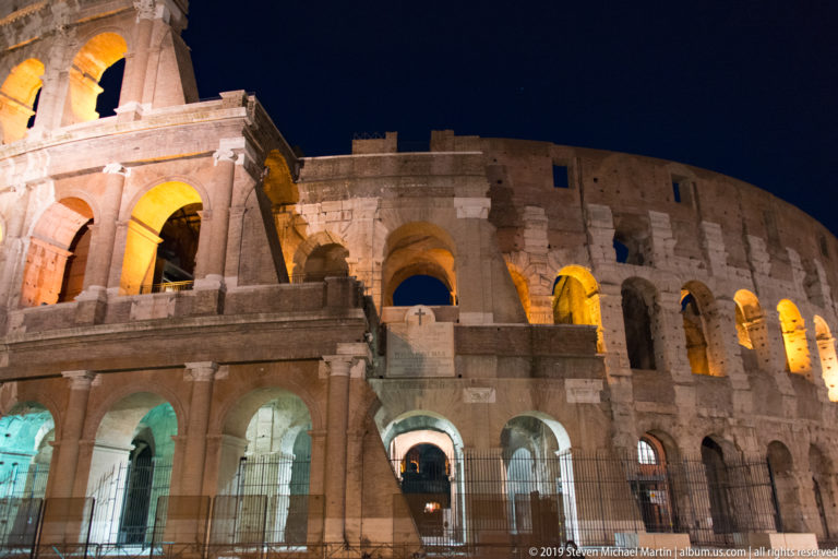 Close night view along the curving Colosseum exterior, with illuminated stacked arches and the surviving upper wall.