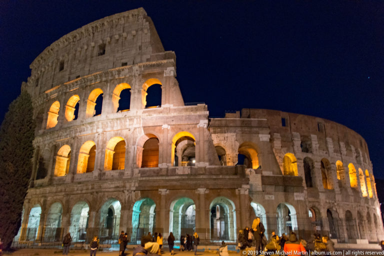 Illuminated Colosseum at night, seen obliquely with glowing arcades, surviving upper wall, and visitors gathered at street level.