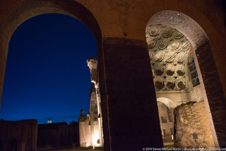 Brick arches framing illuminated Roman ruins, including a coffered vault with circular and polygonal recesses at right.