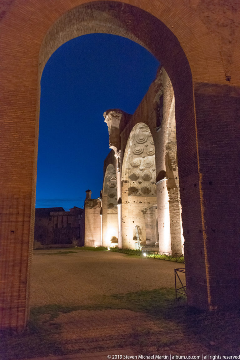 Ancient Forum of Augustus masonry illuminated at night and framed by a large brick arch, with blue sky visible beyond.