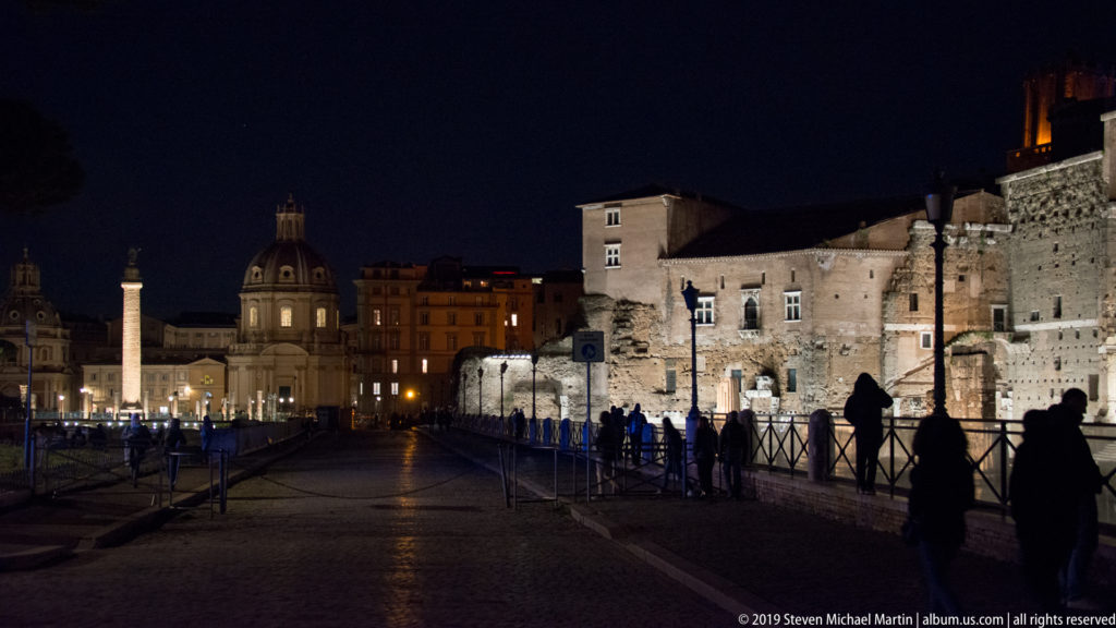 Night walkway beside the Forum of Augustus wall, with pedestrians, Trajan’s Column, and Santa Maria di Loreto visible in the distance.