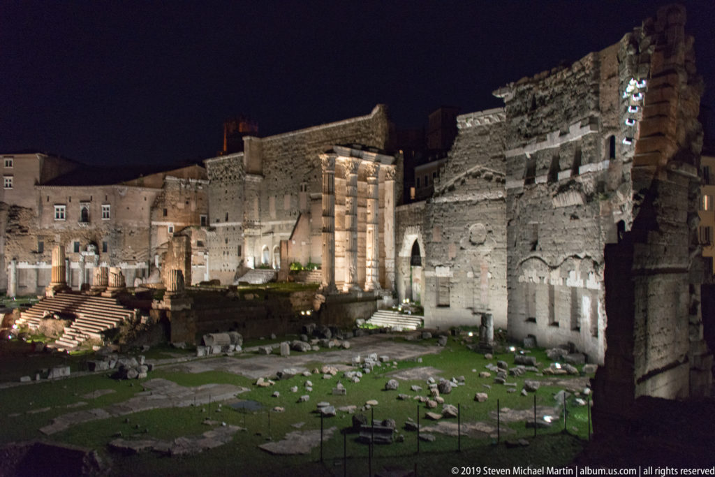 Wide night view of the Forum of Augustus with temple columns, scattered marble blocks, grass, and the high perimeter wall.