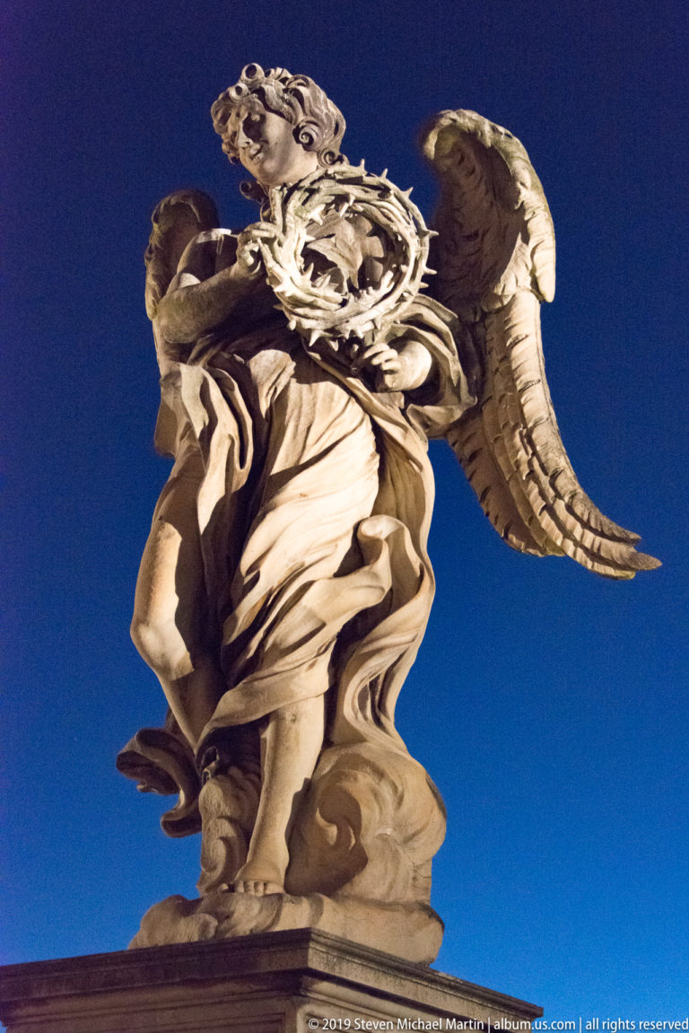Low-angle night view of the Angel with the Crown of Thorns on Ponte Sant’Angelo, holding the circular crown across its body.