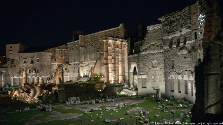 Elevated night view across the Forum of Augustus, with scattered stone remains and the illuminated columns of the Temple of Mars Ultor.