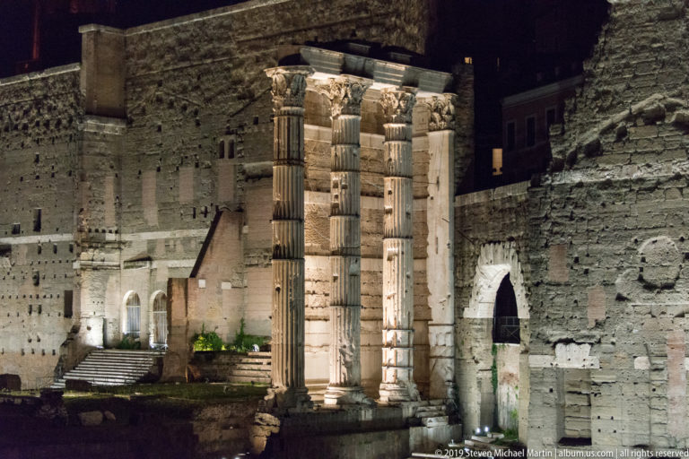 Three floodlit Corinthian columns of the Temple of Mars Ultor standing before the high stone wall of the Forum of Augustus.