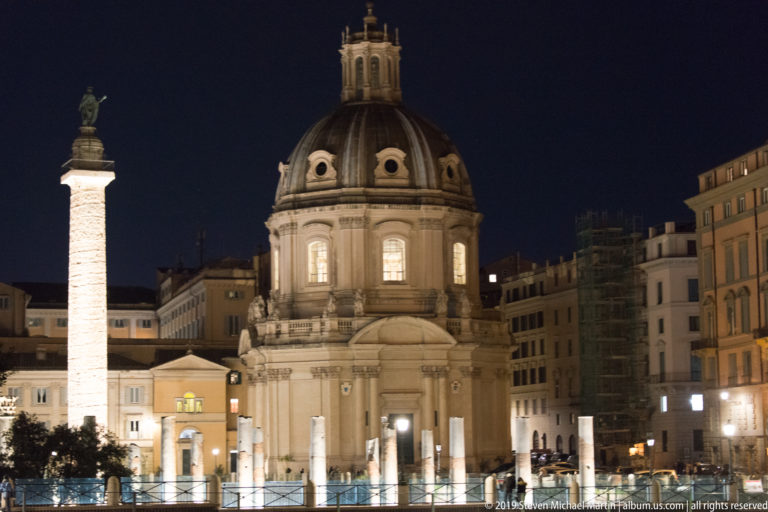 Trajan’s Column beside the illuminated dome of Santa Maria di Loreto at night, with archaeological remains in the foreground.