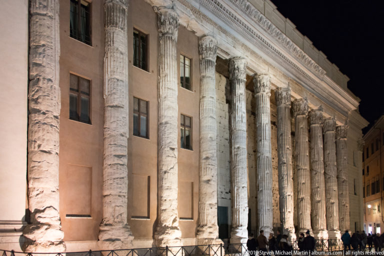 Floodlit Corinthian columns of the Temple of Hadrian incorporated into a building façade at Piazza di Pietra, with pedestrians below.
