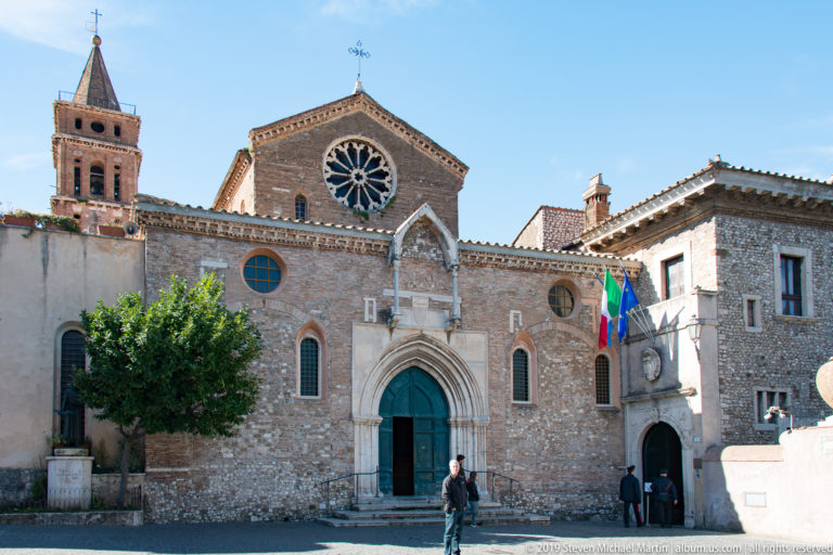 Stone church façade with pointed portal, rose window, brick bell tower, flags, and visitors in a Tivoli square.