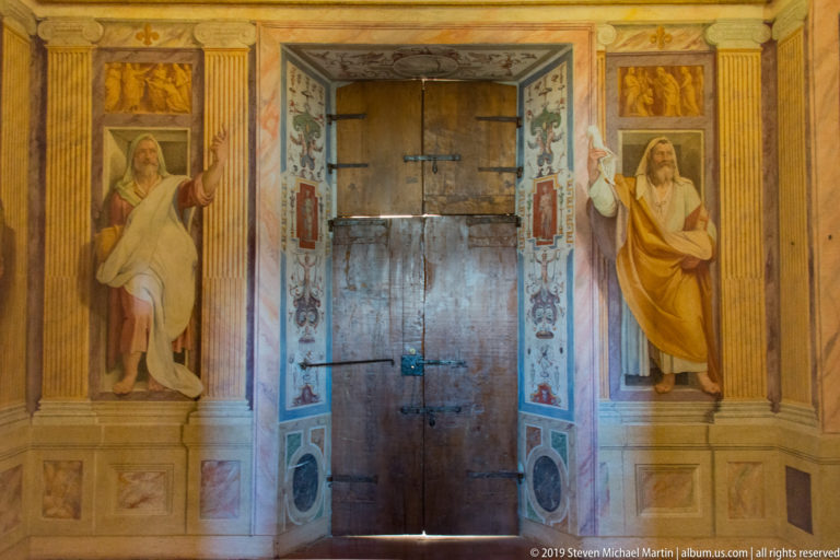Closed wooden door flanked by painted robed figures, fluted pilasters, small panels, and decorative borders.