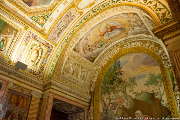 Pastoral landscape fresco framed by an ornate arch covered with painted figures, medallions, and patterned decoration.