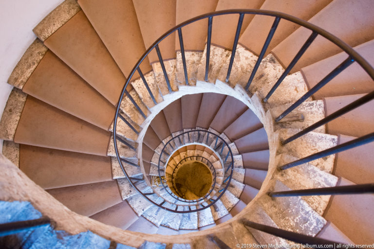Top-down view of a circular stone spiral staircase with dark metal railing winding around an open central well.