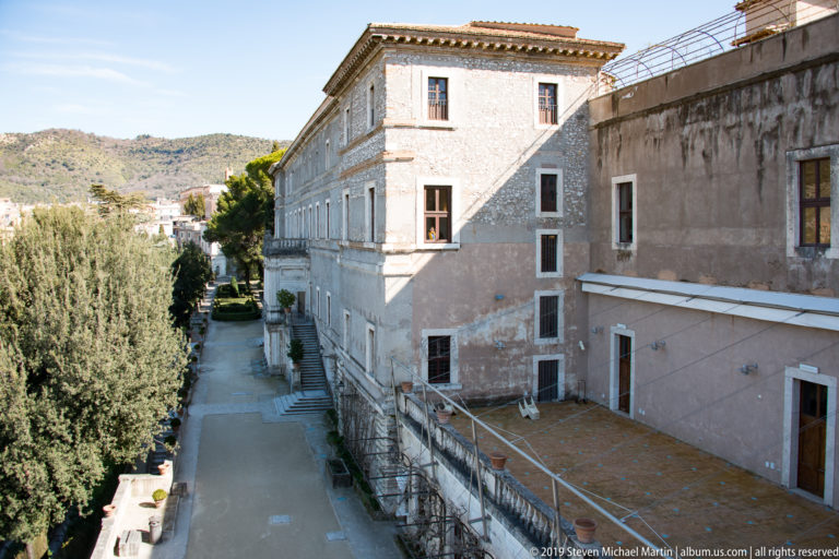 Long weathered palace façade with rows of windows beside a broad terrace passage and trees.