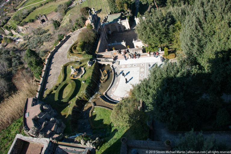 High-angle view of clipped hedges, sculptural fountains, curved basins, paths, and visitors in the Villa d’Este gardens.