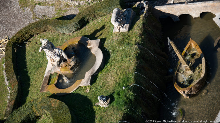 High-angle view of sculptural fountain basins, a standing figure, clipped hedges, and narrow water channels.