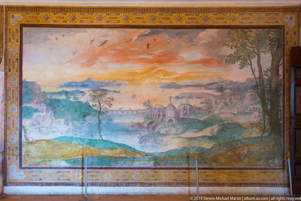 Large framed landscape fresco with water, hills, buildings, bridges, and distant settlements beneath a warm sky.