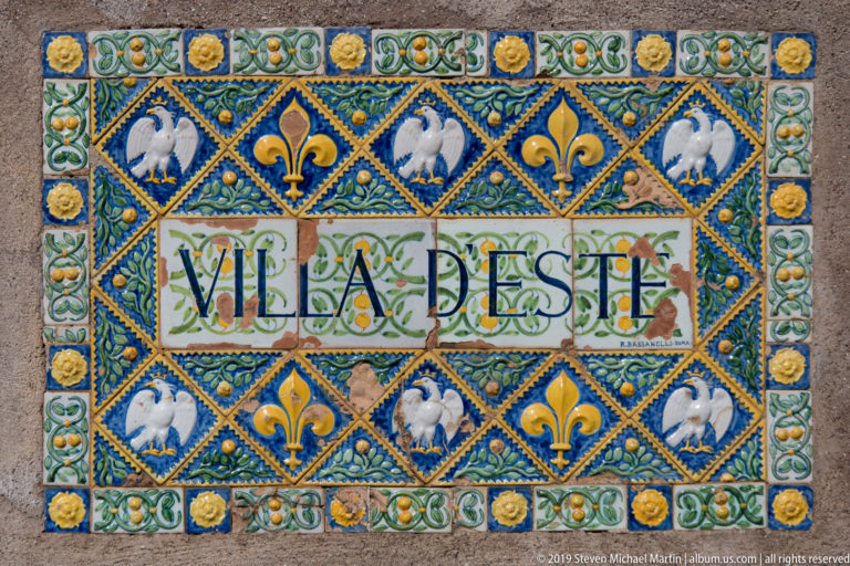 Colorful ceramic plaque reading “VILLA D’ESTE,” bordered by fleur-de-lis, white eagles, rosettes, and blue ornament.