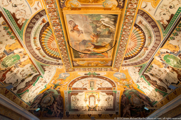 Symmetrical frescoed ceiling with central mythological panel, fan-shaped compartments, heraldry, and colorful grotesque ornament.