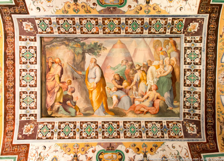Framed fresco of a bearded standing figure addressing seated and kneeling people before a mountainous landscape.