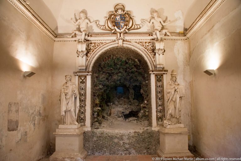 Rock-lined arched doorway framed by two standing statues, decorative columns, and a heraldic cartouche.