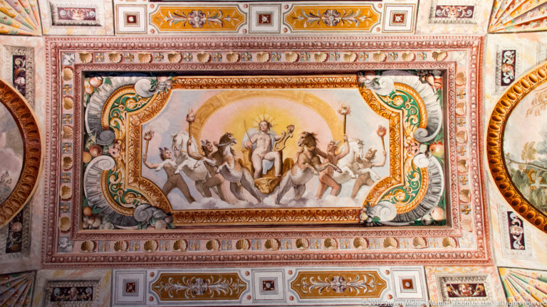 Classical figure and lively procession painted in a ceiling panel surrounded by colorful Renaissance ornament.