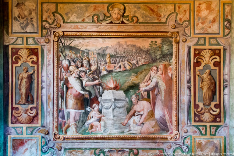 Fresco of armed and robed figures gathered around a flaming altar with a distant landscape behind.