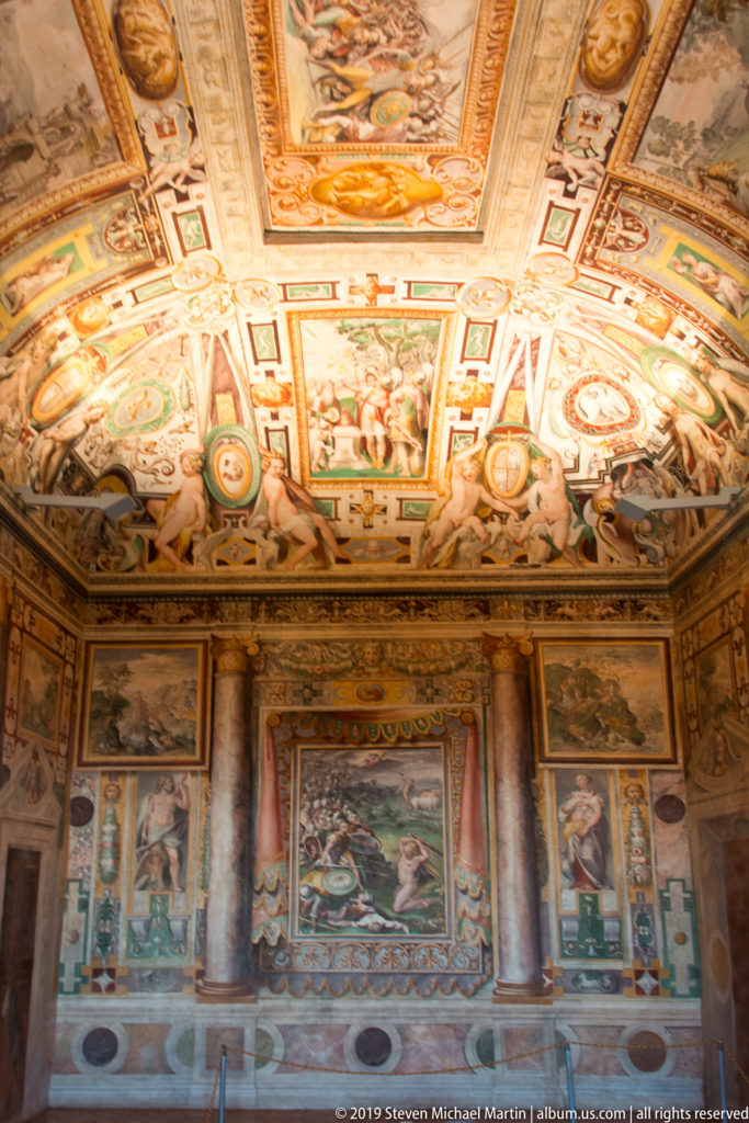 Richly frescoed room with decorated vault, painted walls, two columns, and multiple framed narrative scenes.