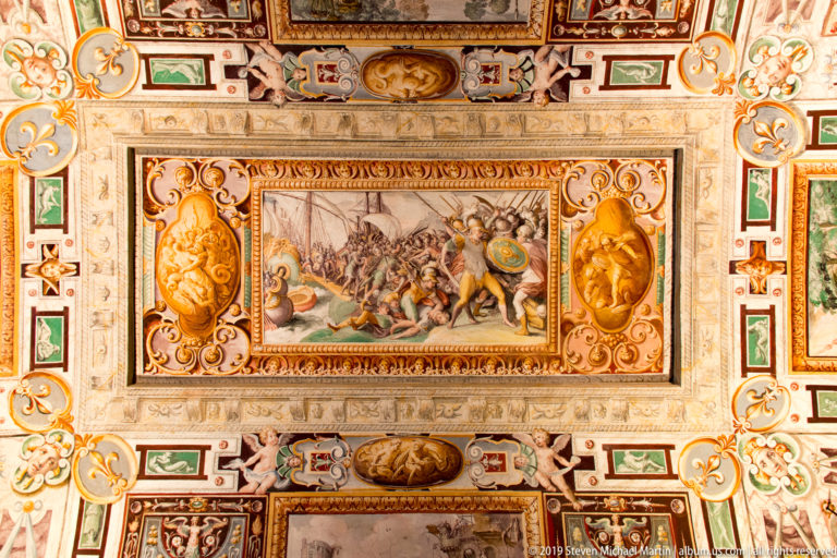 Rectangular mythological battle scene surrounded by dense painted grotesques, masks, medallions, and scrolling ceiling ornament.