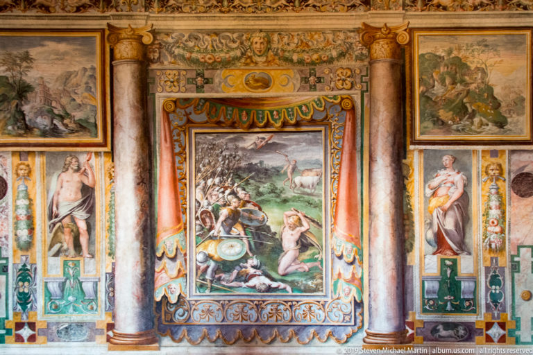 Central mythological fresco framed by painted columns, standing figures, garlands, and smaller narrative panels.