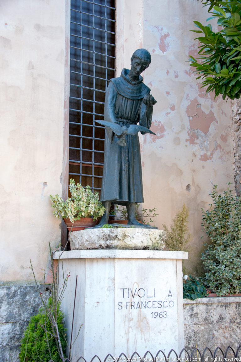 Dark statue of a Franciscan friar on a pedestal inscribed “TIVOLI S. FRANCESCO 1963,” beside plants and a weathered wall.