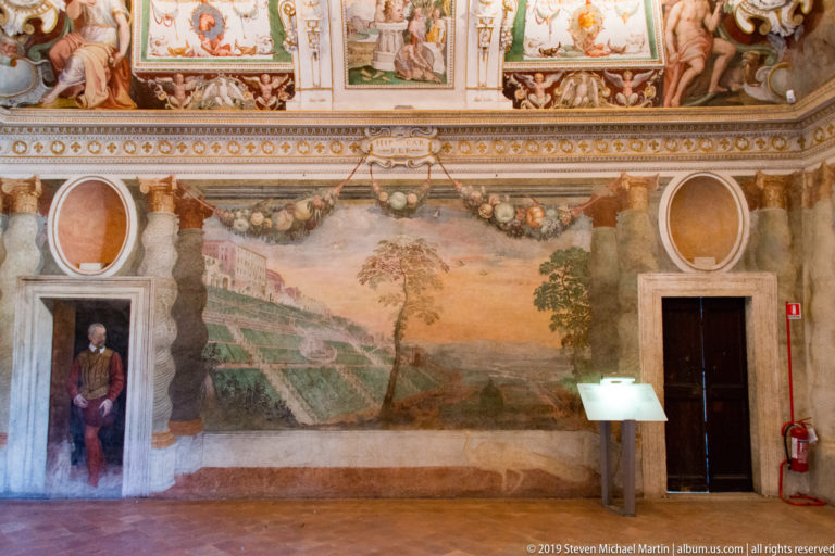 Frescoed room wall with large landscape scene, oval panels, painted ornament, doorway, and interpretive stand.