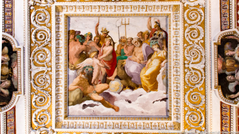 Crowded mythological fresco of classical figures within an ornate white-and-gold rectangular ceiling frame.
