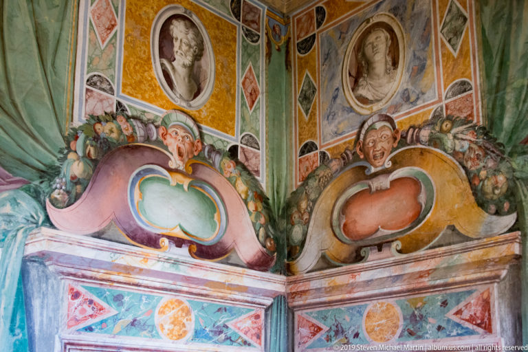 Colorful frescoed corner with portrait medallions, fruit garlands, masks, geometric panels, and scrolling cartouches.