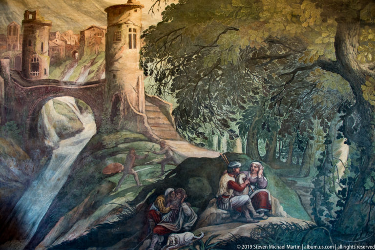 Interior fresco of a wooded landscape with stream, bridge, buildings, and small groups of resting figures.