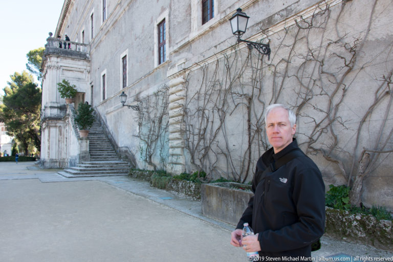 Man in black jacket holding a bottle beside a pale palace wall with bare vines and stone staircase.