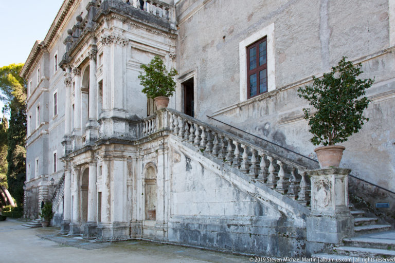 Stone staircase with balustrade climbs along a weathered palace façade with tall windows and potted plants.