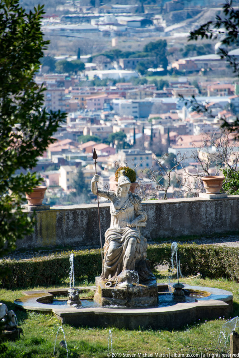 Classical figure in a circular fountain basin with small jets and Tivoli spread across the distant valley.
