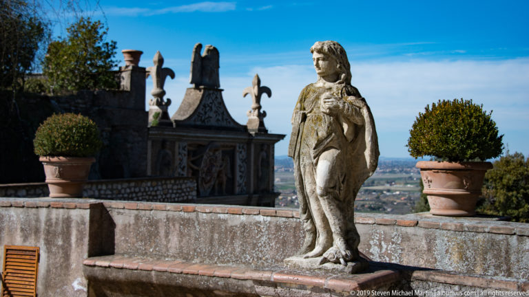 Weathered standing female statue between potted shrubs on a terrace overlooking the distant landscape.