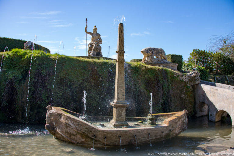 Stone boat-shaped fountain with tall central mast and small jets in a pool below an ivy-covered terrace.
