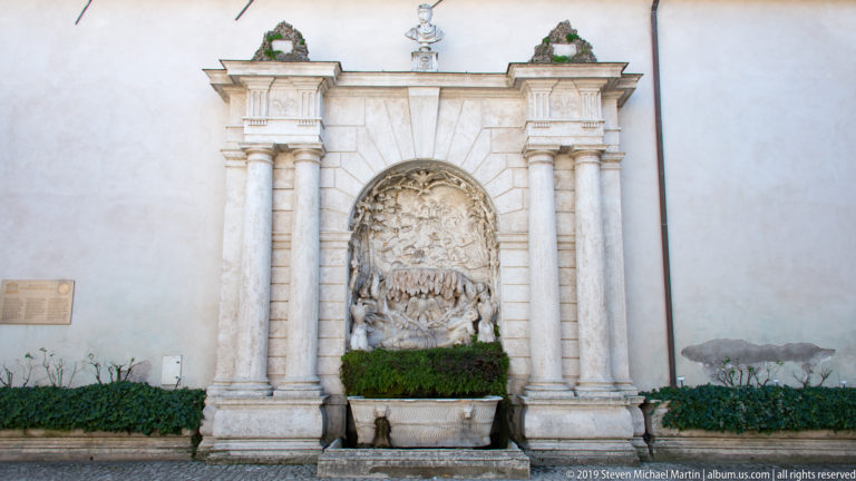 Monumental landscape relief framed by paired columns and entablatures above a planted rectangular fountain basin.