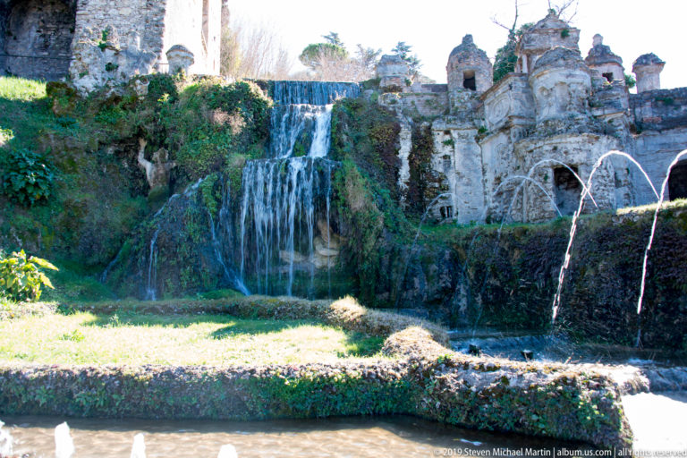 Broad cascade beside ornate weathered grotto architecture, with slender fountain jets crossing the foreground.
