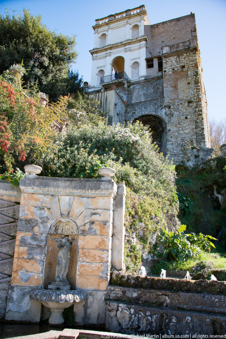 Small niche fountain below a planted slope and pale garden structure built into a steep terrace.