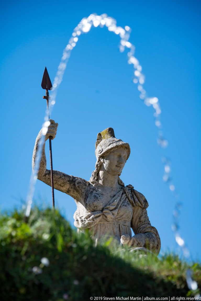 Pale classical fountain figure holding a staff beneath a high arc of water against blue sky.
