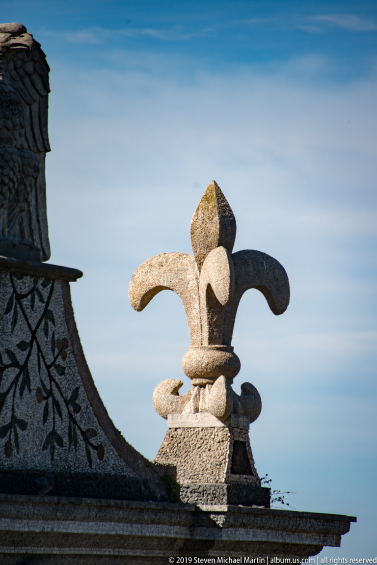 Weathered stone fleur-de-lis finial silhouetted against a pale blue sky beside patterned masonry.