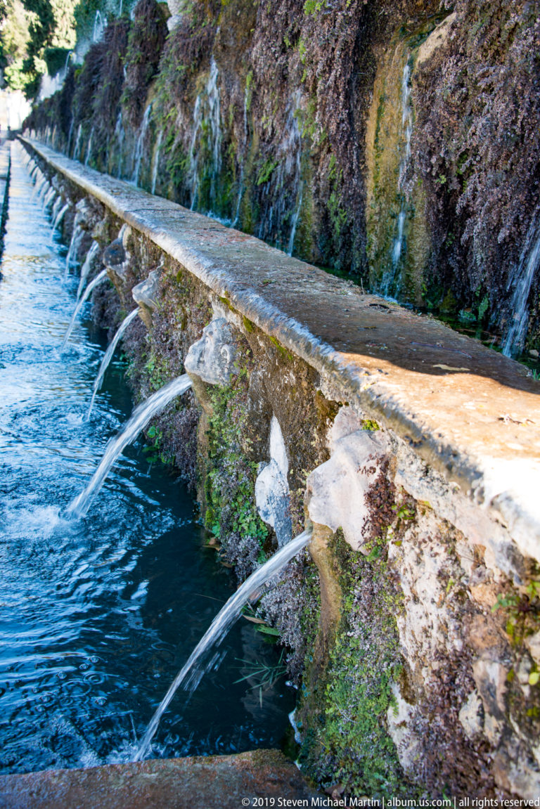Parallel water streams issue from repeated carved spouts along a mossy wall and blue-green channel.