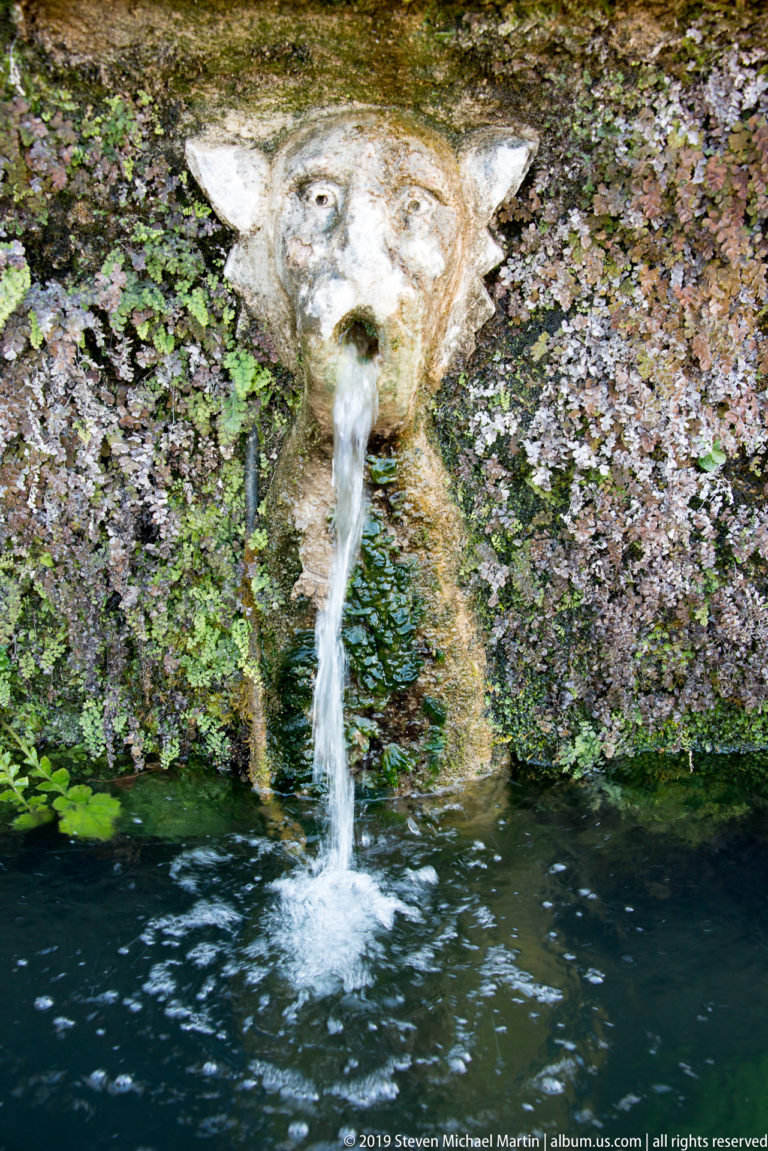 Weathered carved face spouting water from a mossy wall into a narrow channel.