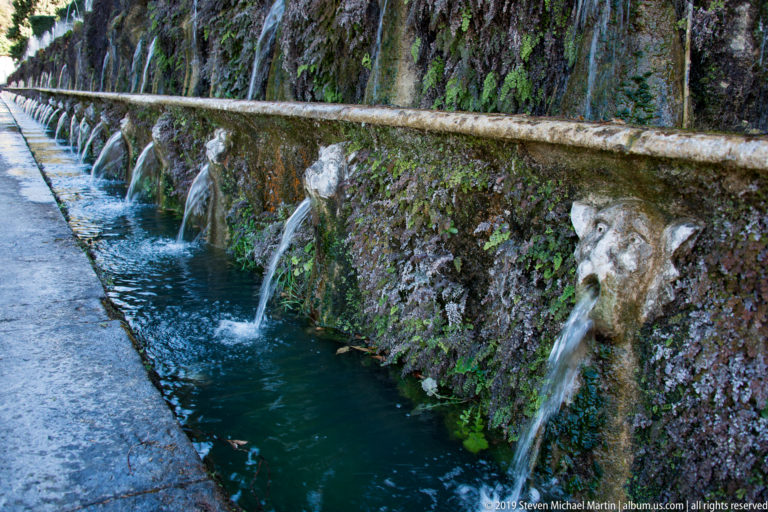 Row of carved masks spouting water into a long narrow channel along a moss-covered wall.